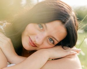 Young woman with dark hair and green eyes resting her chin on her hands, smiling softly in warm sunlight.