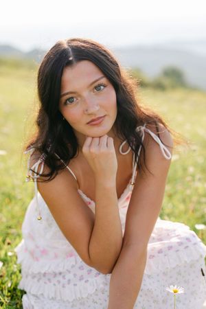 Young woman in white floral dress sitting in a sunny green meadow with daisies, posing with hand near chin.