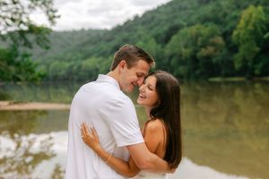 Happy couple embracing and smiling by a serene river with lush green hills in the background.