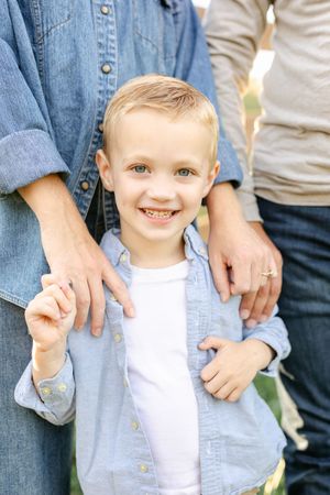 Smiling blonde boy in a blue shirt stands between two adults outdoors during a family photo session.