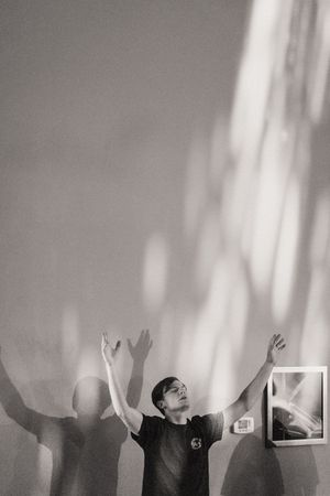 Black and white photo of a young man with arms raised, casting dramatic shadows on a wall with light streaming in.