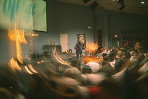 Speaker presenting on stage to a large audience in a modern church or conference hall with warm lighting.