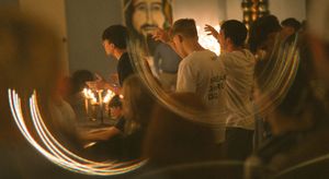 People lighting candles in a dimly lit church with a portrait of Jesus Christ on the wall during a religious ceremony.