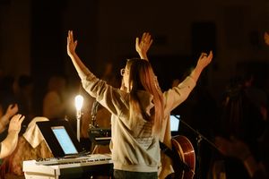 Woman raising hands in worship on stage with keyboard and guitar at a dimly lit church concert.