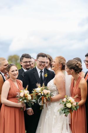 Bride and groom smile with wedding party, bridesmaids in rust dresses holding bouquets, groomsmen in black suits outdoors.