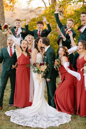 Wedding party celebrates as bride and groom kiss, bridesmaids in red gowns and groomsmen in dark green suits cheering outdoors.