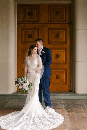 Bride in lace gown holding bouquet stands with groom in navy suit before ornate wooden doors on wedding day.