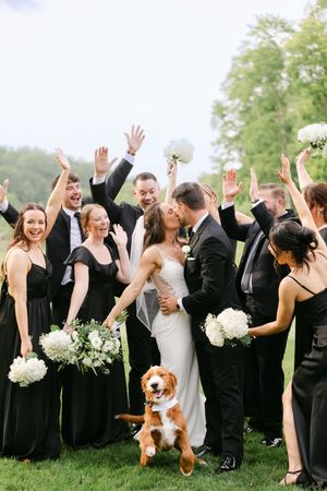 Wedding party in black attire cheering as bride and groom kiss, with an adorable dog wearing a bow tie in the foreground.