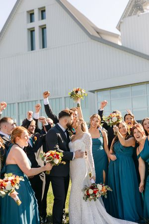 Bride and groom kiss surrounded by cheering wedding party in teal dresses outside a white barn venue.