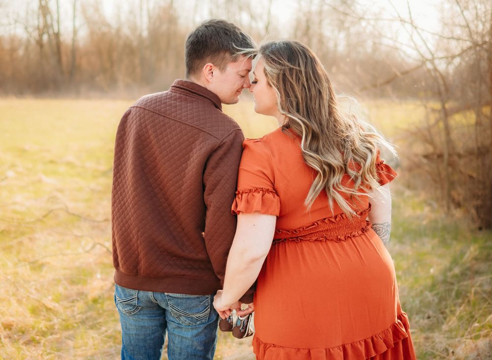 Expectant couple in rustic attire sharing an intimate moment in a golden field at sunset.