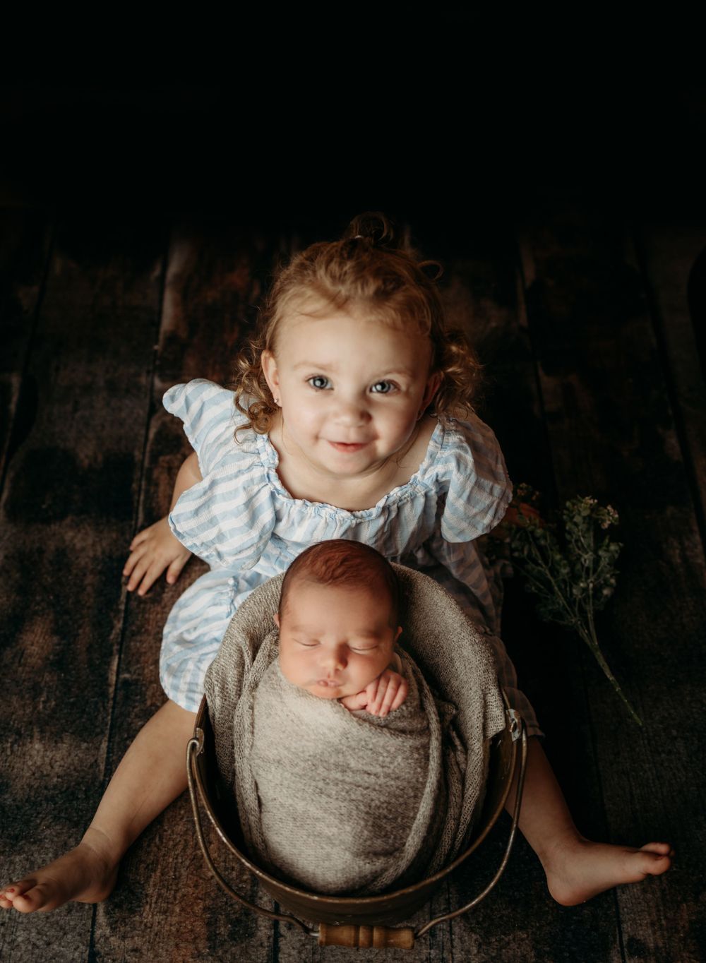 Two siblings posing together with one seated in a rustic basket against a dark background.