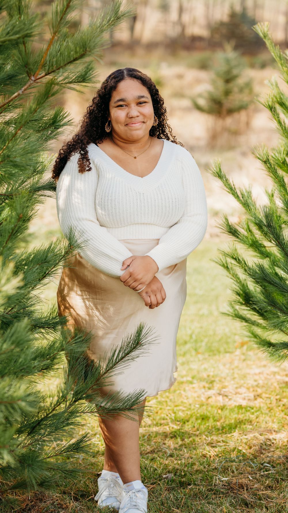 A person in a white sweater and beige skirt poses between evergreen trees in an outdoor portrait setting.