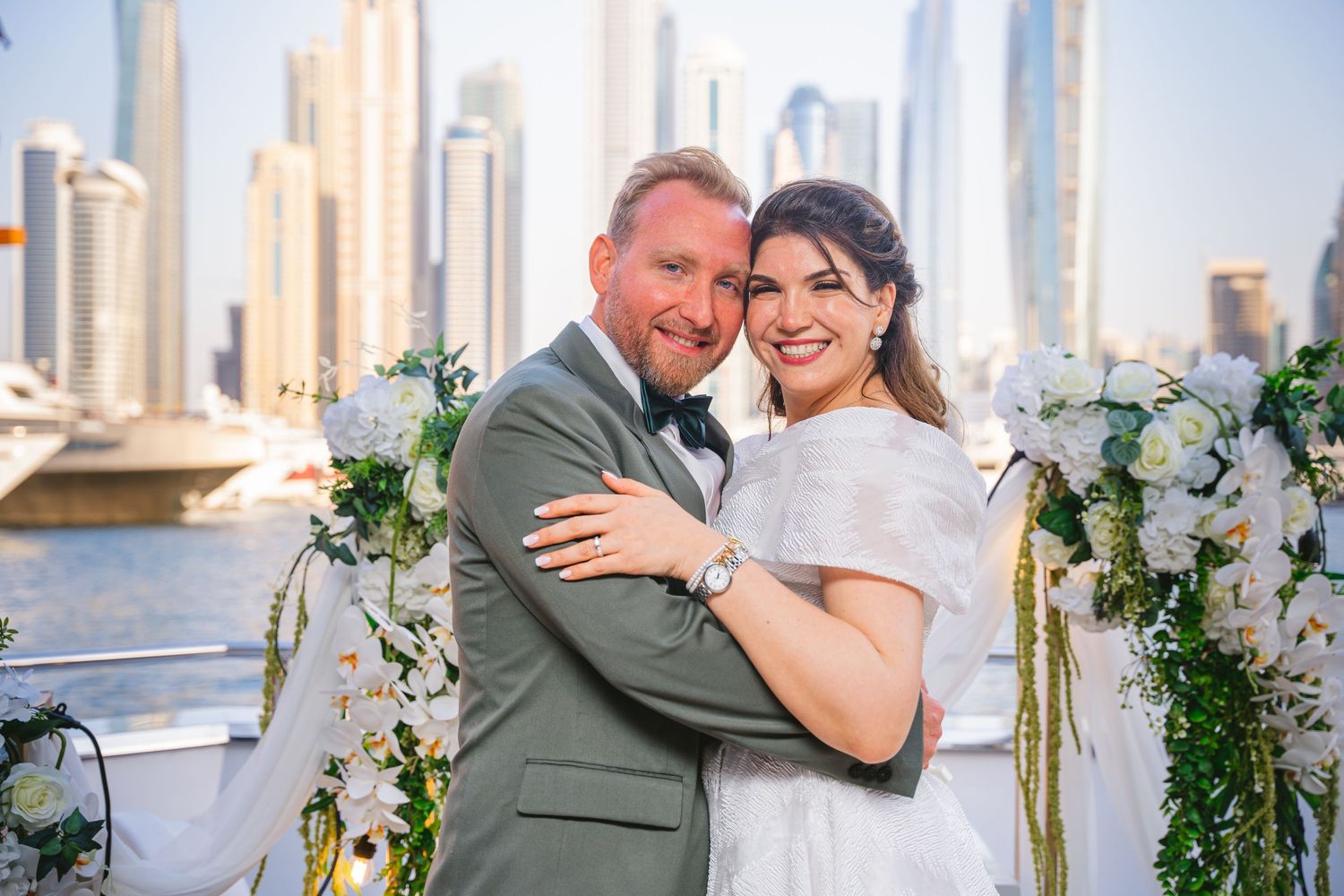A newlywed couple poses against Dubai's modern skyline with white floral decorations.