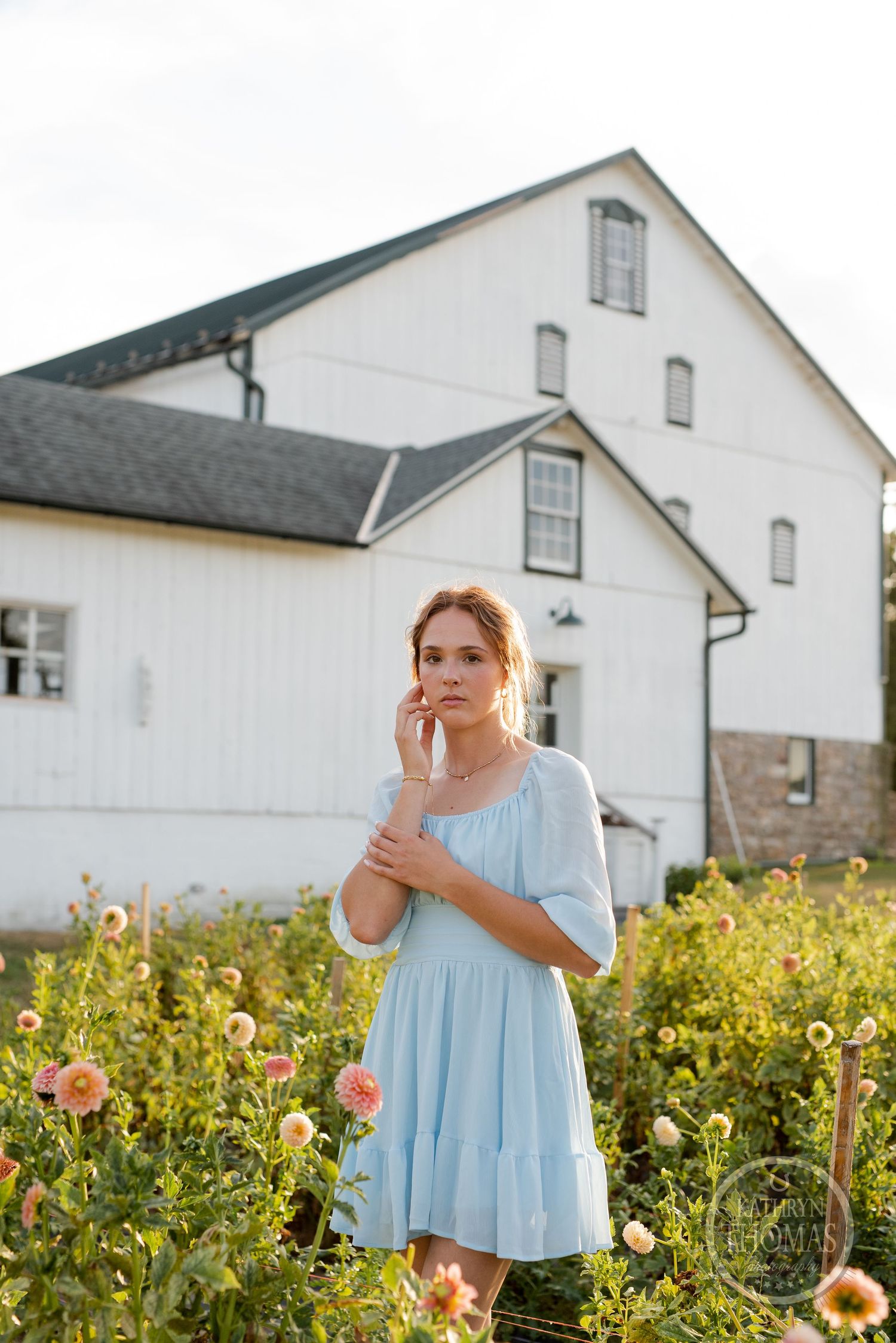 Light blue dress stands out among blooming garden flowers against rustic white barn backdrop.
