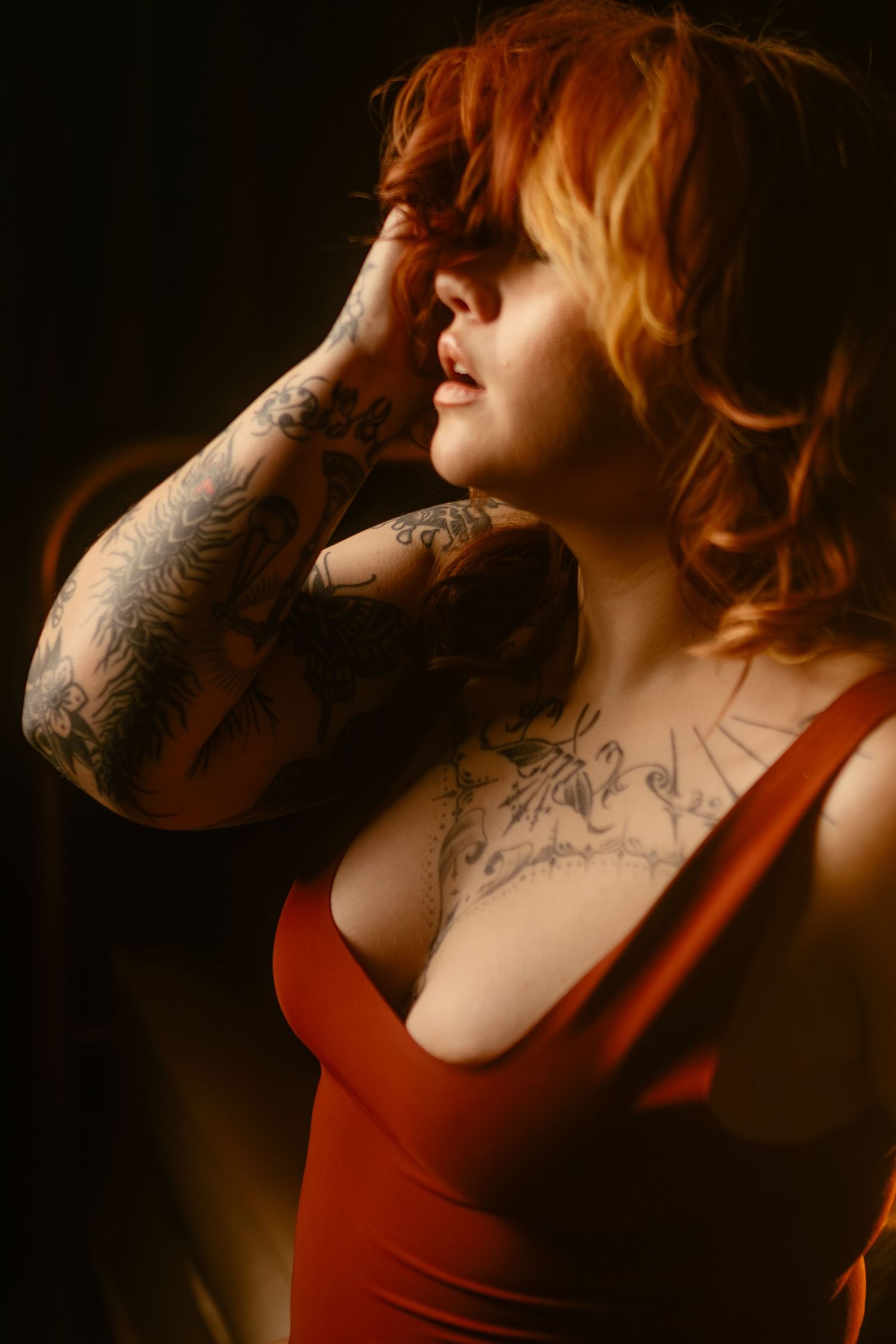 A portrait in warm light shows a red-haired figure in a red tank top with intricate arm and chest tattoos in DFW boudoir session.