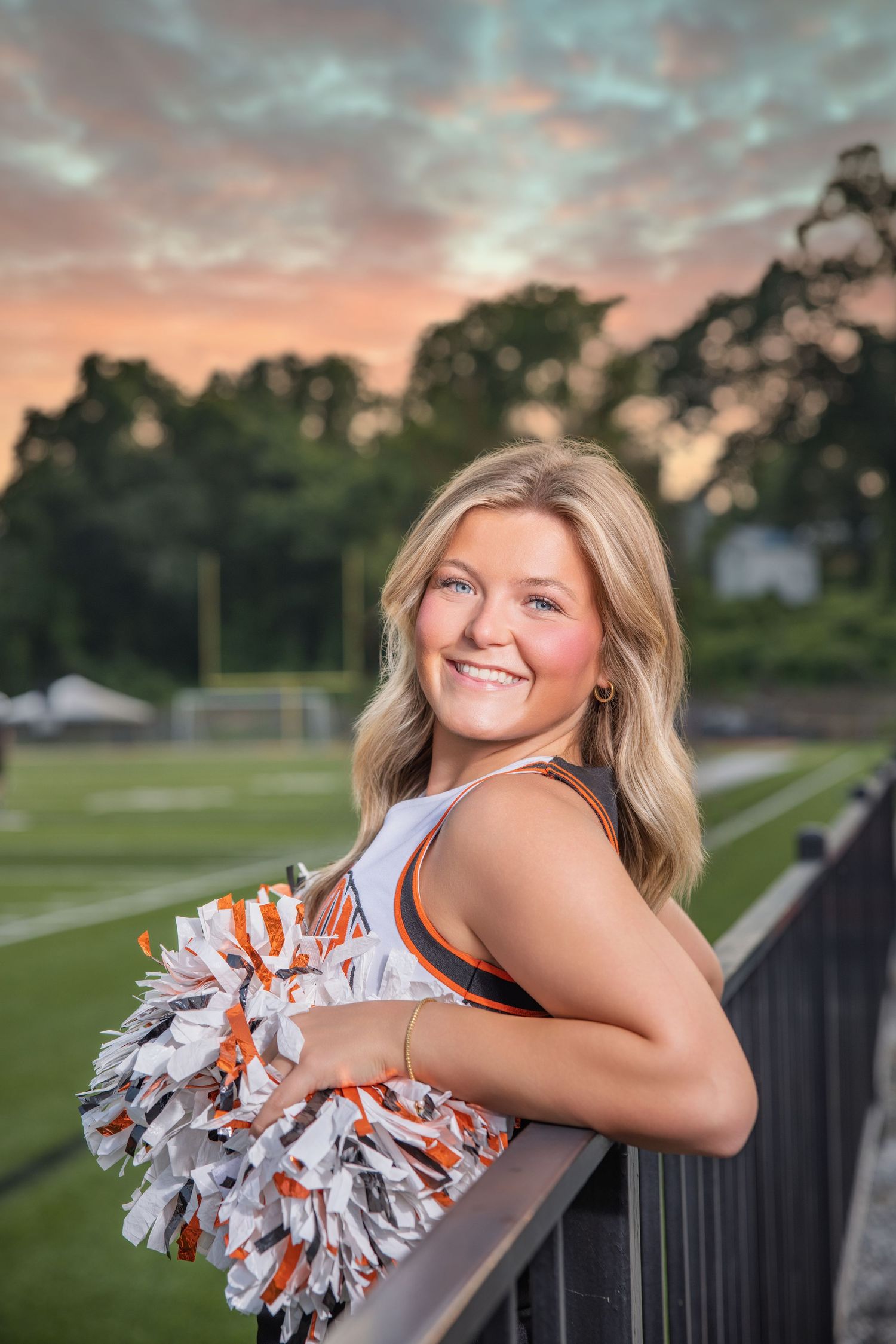 A cheerleader in white and orange uniform holds pom-poms while leaning on a railing at sunset with trees in background.