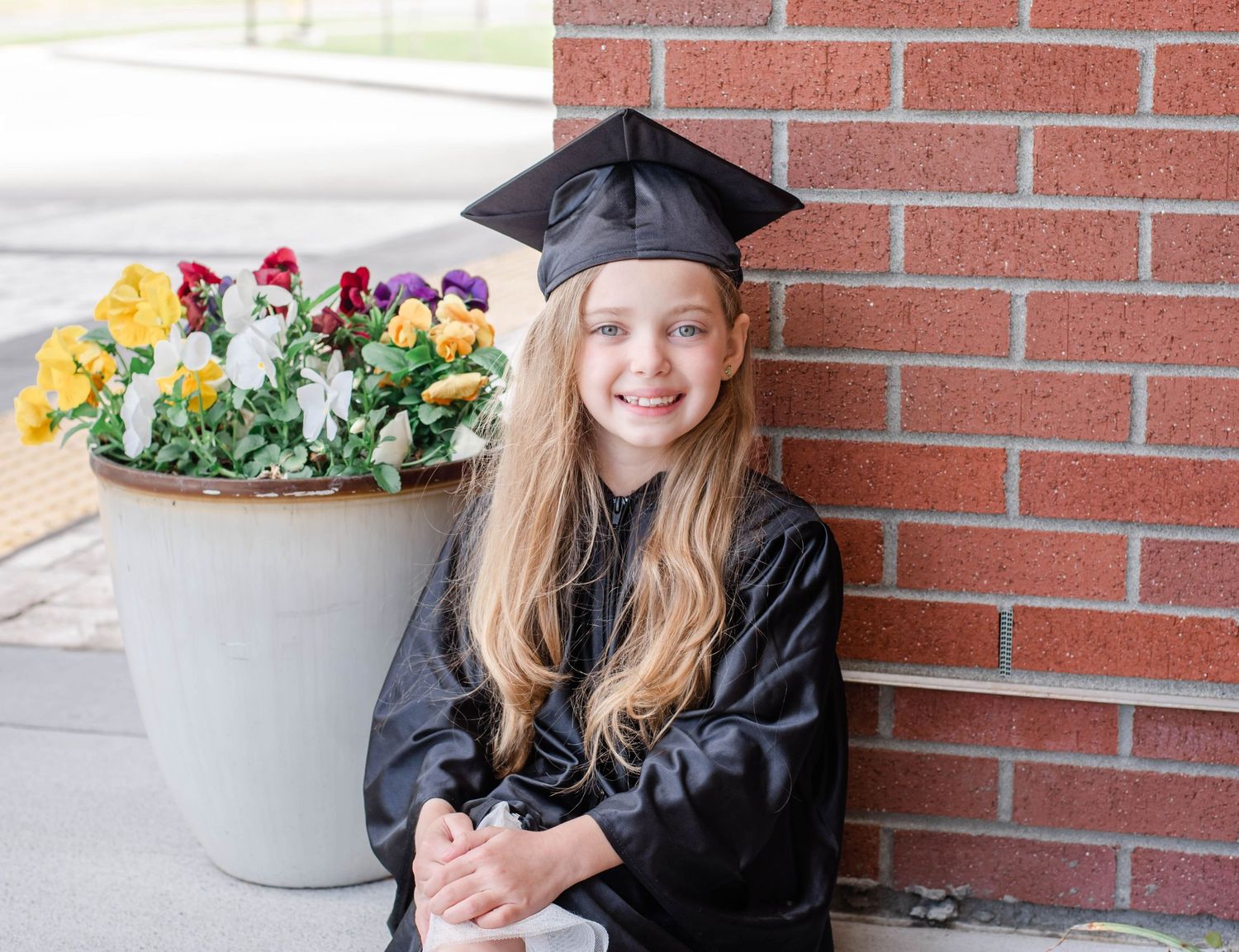 Graduate poses near decorative flower planter against red brick wall for graduation portraits.