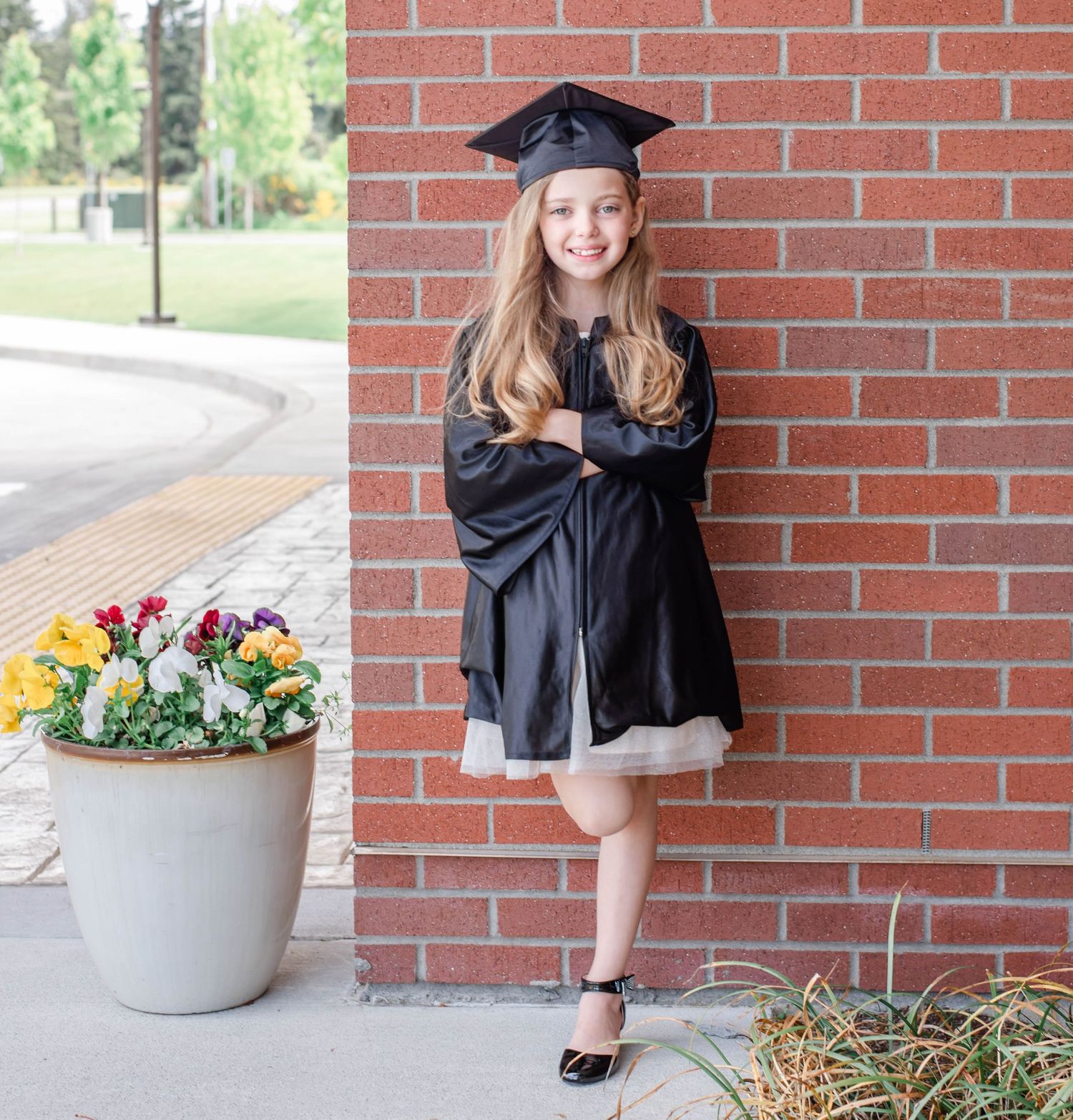 Graduate wearing black cap and gown sits beside floral planter during outdoor photo session.