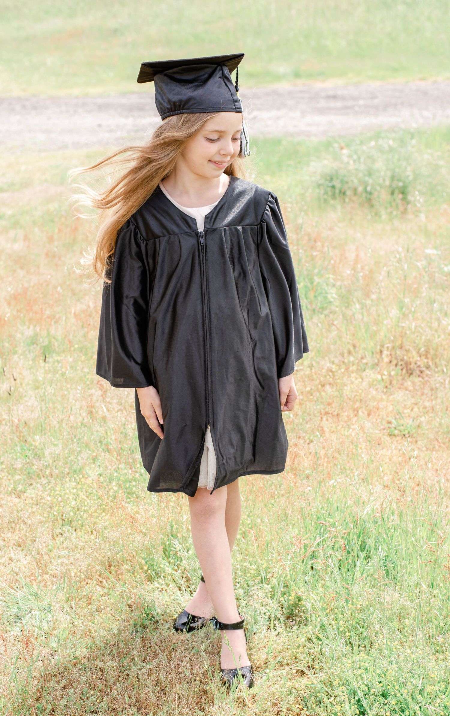 Graduate in flowing black gown walks through sunny field during photo session.