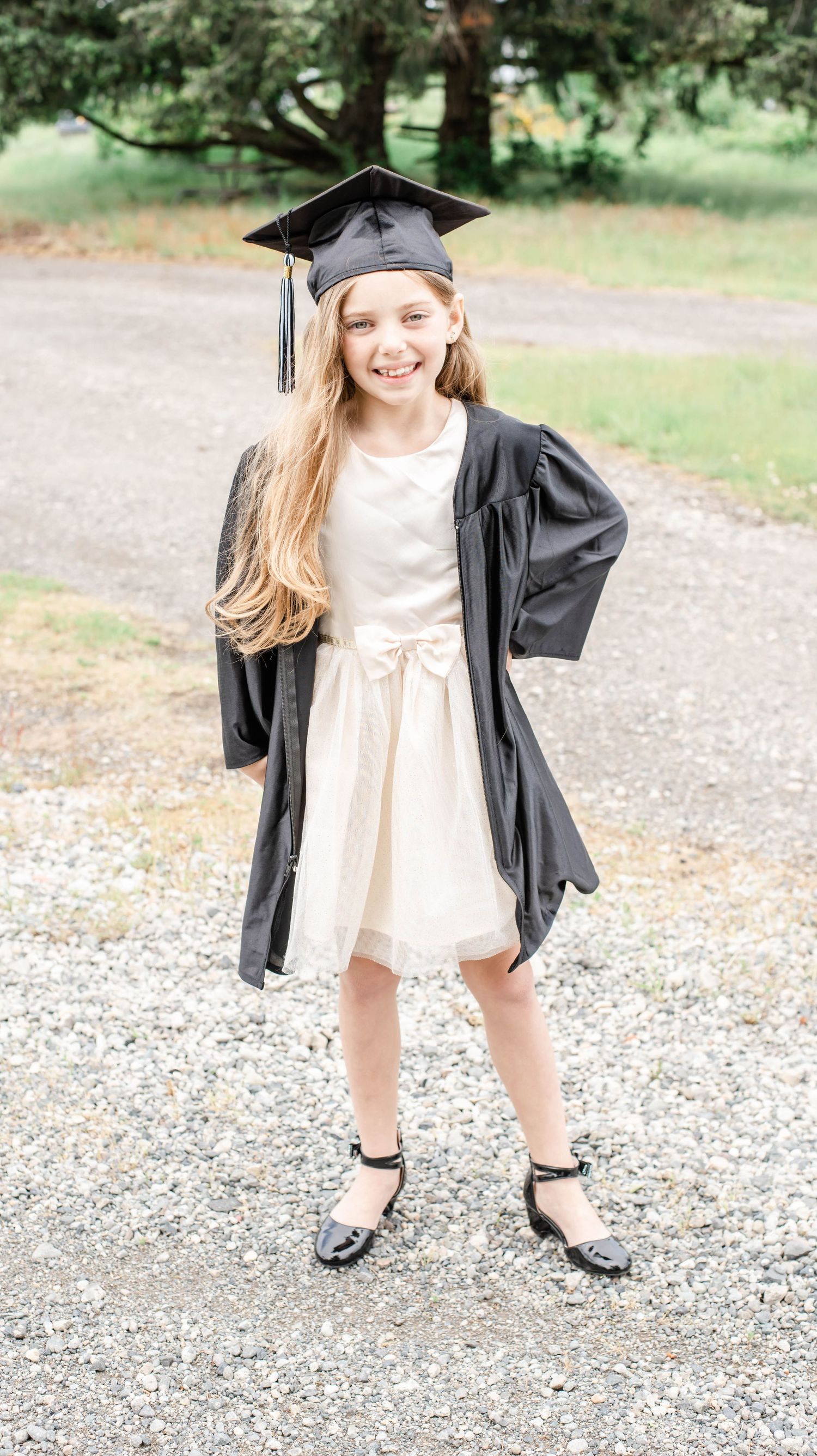 Graduate in white dress and black cap and gown poses playfully on gravel path.