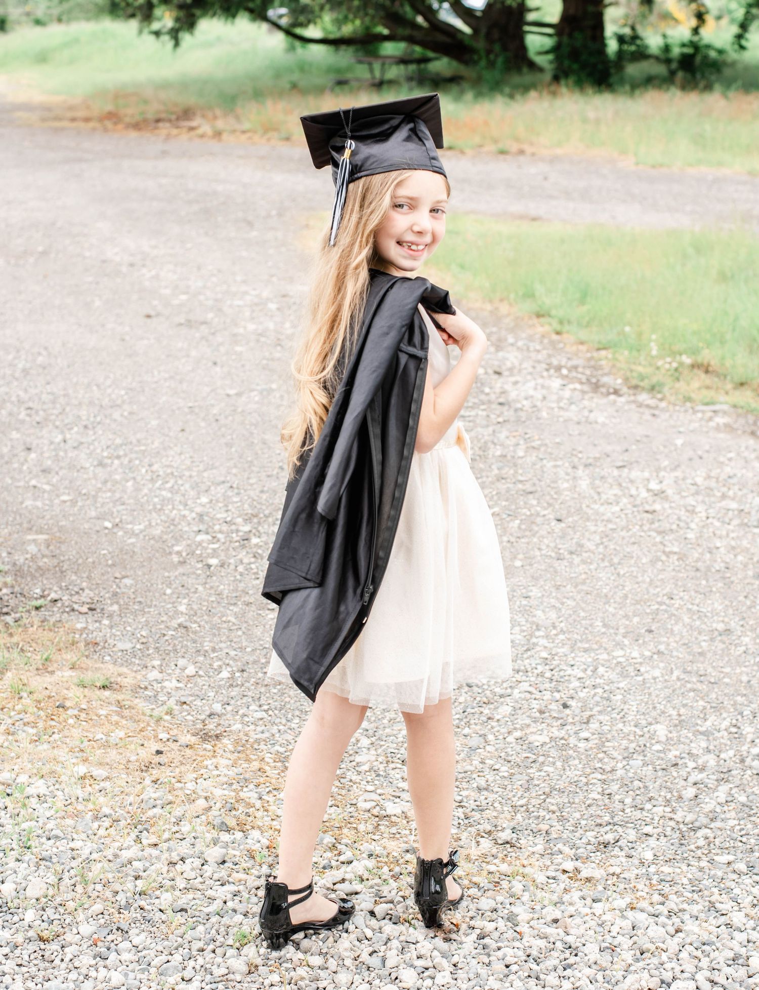 Graduate in flowing white dress and black academic regalia twirls during outdoor photo shoot.