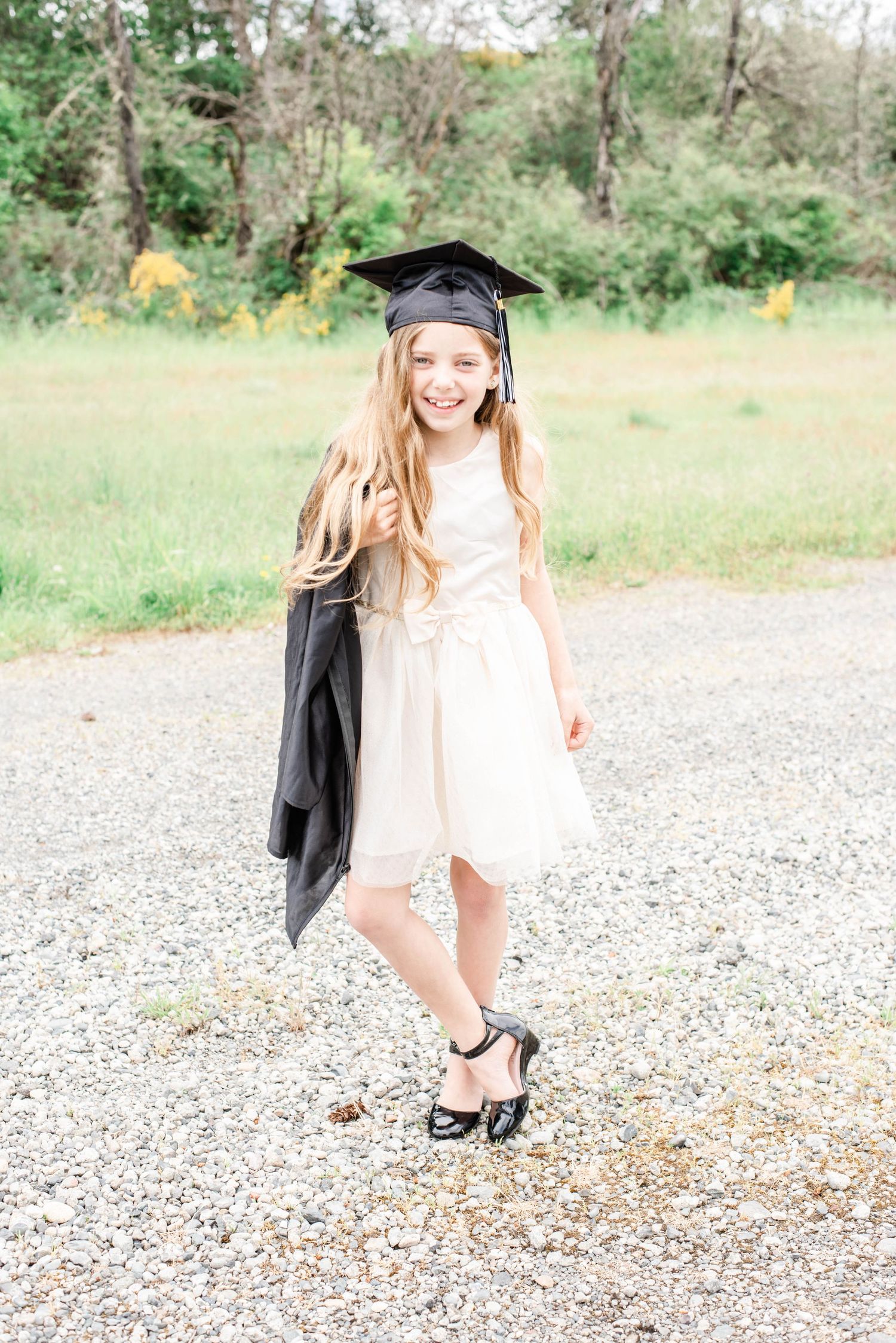 A graduate in a white dress and heels poses playfully on a gravel path surrounded by greenery.