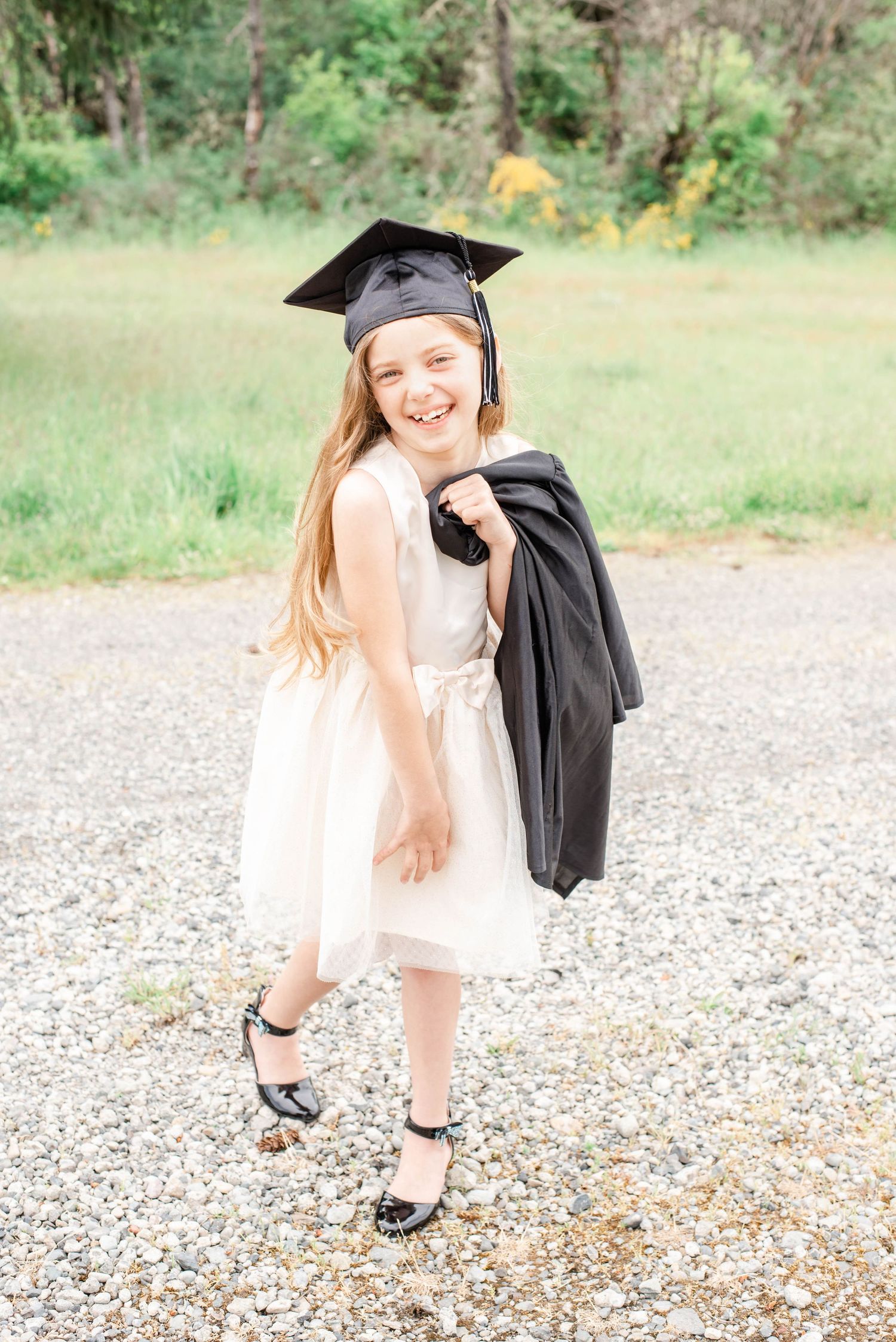Graduate in white dress and black cap poses casually on gravel path with natural backdrop.
