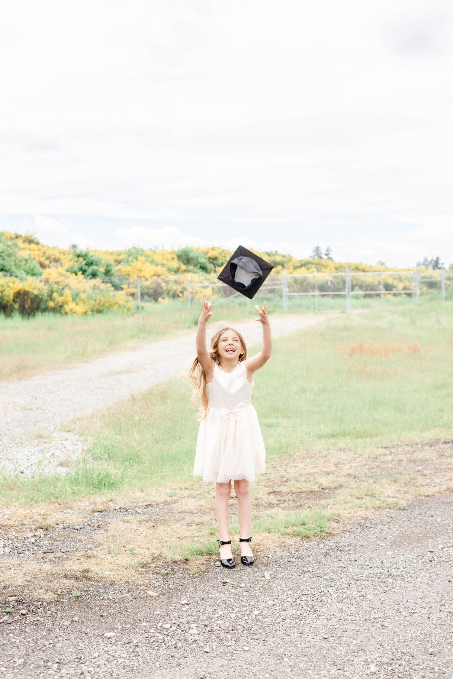 Graduate celebrates on gravel path with cap toss against backdrop of yellow wildflowers.