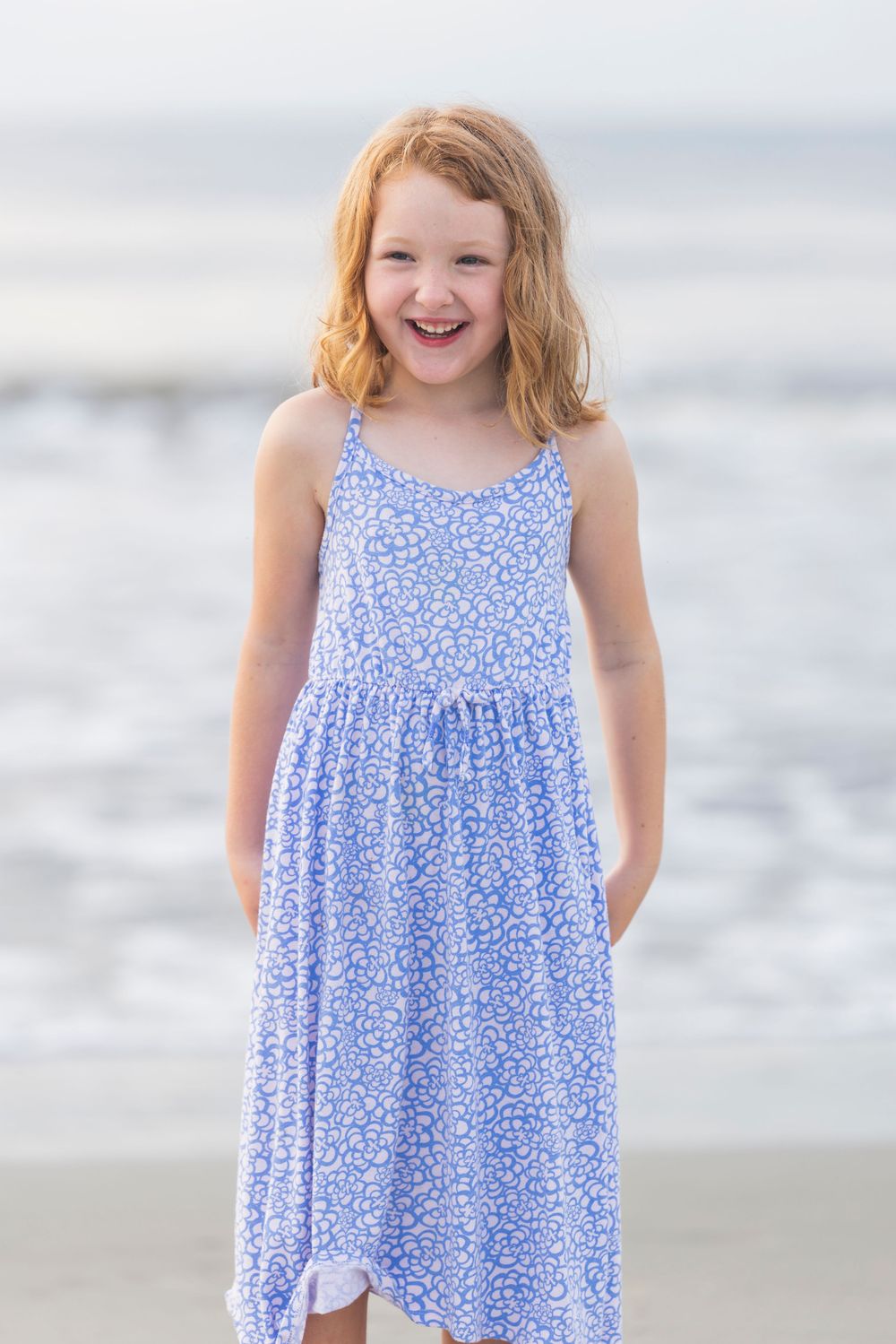 A young child in a blue floral sundress stands joyfully on a beach with waves in the background.
