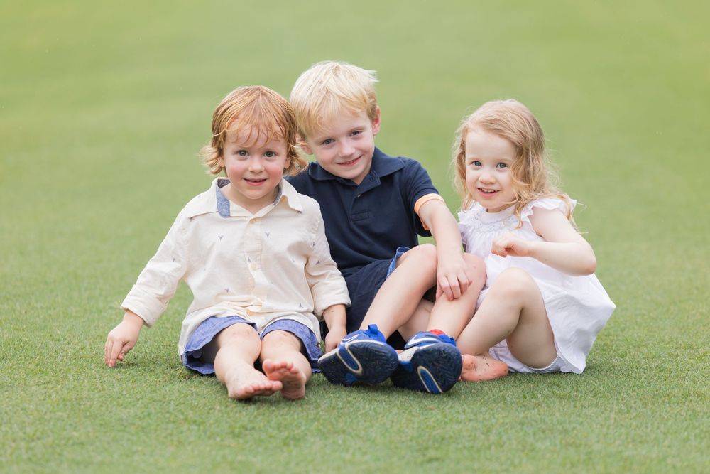 Children sit and play together on a lush green grass field in casual summer clothing.