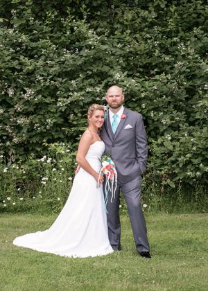 Bride in white gown and groom in gray suit pose together outdoors on their wedding day with lush green foliage behind them.