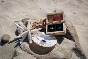 Wedding rings displayed in a wooden box on sandy beach with starfish, driftwood, and seashells.