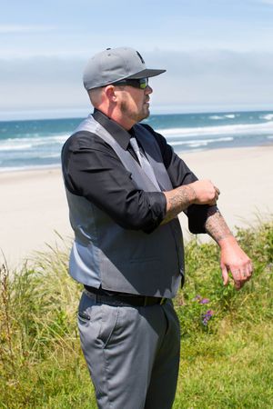 Tattooed man in gray suit, vest, cap, and sunglasses stands on a beach with ocean waves in the background.