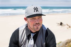 Man in NY Yankees cap and gray jacket smiling on a sunny beach with ocean waves in the background.