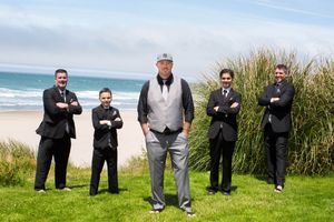 Five men in formal suits pose with arms crossed on a grassy coastal area with ocean waves and sandy beach behind them.