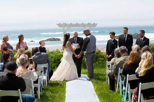Outdoor beach wedding ceremony with couple at altar, officiant, groomsmen, and seated guests on ocean bluff.