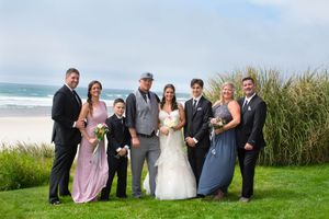 Wedding party poses on green lawn with ocean backdrop, bride in white gown flanked by bridesmaids in pink and blue dresses.
