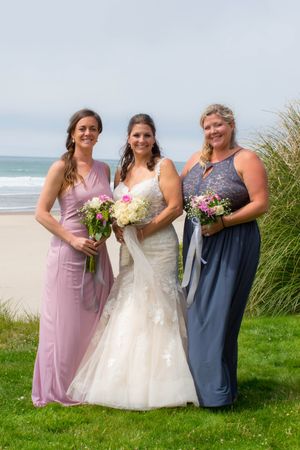 Bride in lace mermaid gown stands with two bridesmaids in pink and blue dresses holding bouquets by beach.