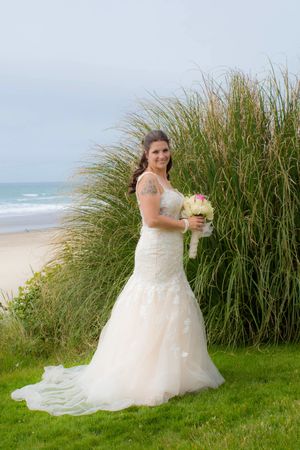 Bride in white lace mermaid gown holding bouquet stands before tall beach grass with ocean view.