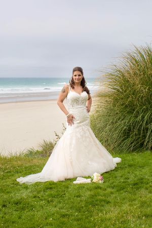 Bride in a fitted lace mermaid wedding gown poses on green grass with a sandy beach and ocean backdrop.