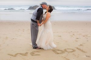 Newlywed couple kissing on beach with WE DO written in sand, bride in white gown, groom in gray suit.