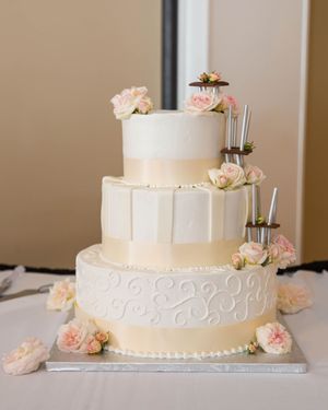 Elegant three-tier white wedding cake adorned with pink roses and decorative scrollwork on silver stand.