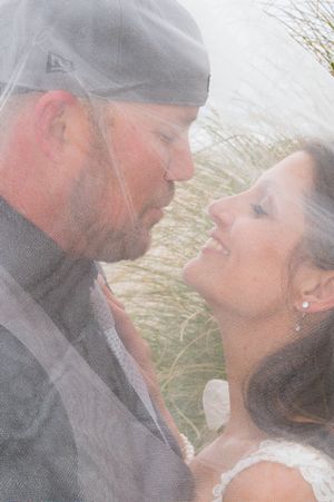 Bride and groom share a romantic almost-kiss beneath a wedding veil outdoors near tall grass.