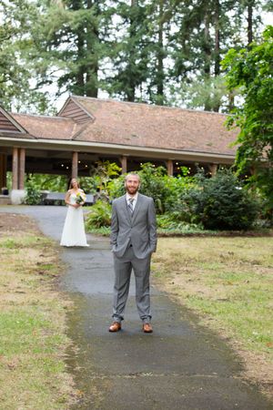 Groom in gray suit stands on garden path with bride in white dress approaching a rustic venue surrounded by tall trees.