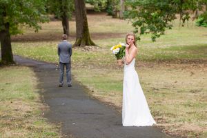 Bride in white gown holds yellow and white bouquet while groom stands on park path in first look wedding moment.