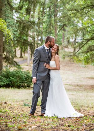 A groom in a gray suit kisses his bride in a white gown outdoors among tall green trees at a wedding.