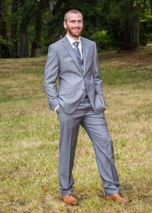 Man in gray suit with blue tie and brown shoes standing on grass with trees in background.