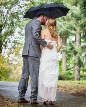 Bride and groom sharing a kiss under a black umbrella in the rain, bride wearing white gown with sunflower hair accessory.