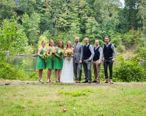 Wedding party posing outdoors near a pond, bride in white, bridesmaids in green dresses, groomsmen in suits.