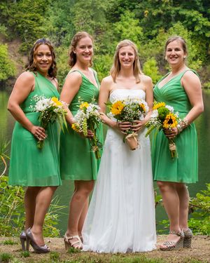 Bride in white gown with three bridesmaids in green dresses holding sunflower bouquets outdoors near a lake.